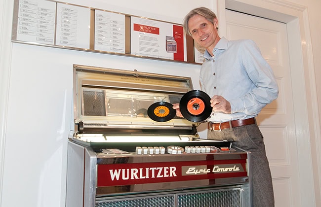 Mann hält Schallplatten neben einer Wurlitzer-Jukebox in einem gut beleuchteten Raum mit weißem Hintergrund.