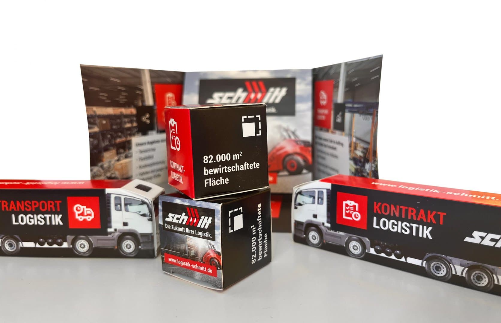 Zwei kleine Logistik-Modell-LKWs und zwei kleine Kisten mit Logistik-Branding vor einem gefalteten Hintergrunddisplay.