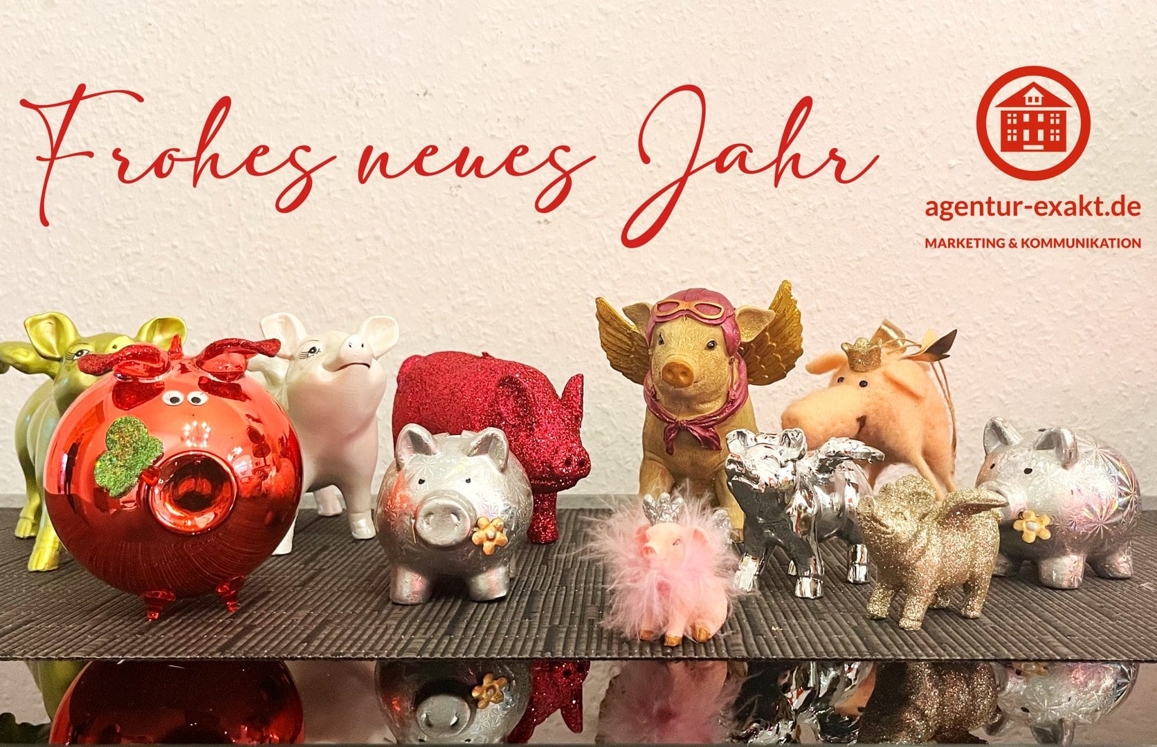Kleine Deko-Schweine und ein Plüschschwein auf einem Tisch mit „Frohes neues Jahr“-Schriftzug und „agentur-exakt.de“-Logo im Hintergrund.