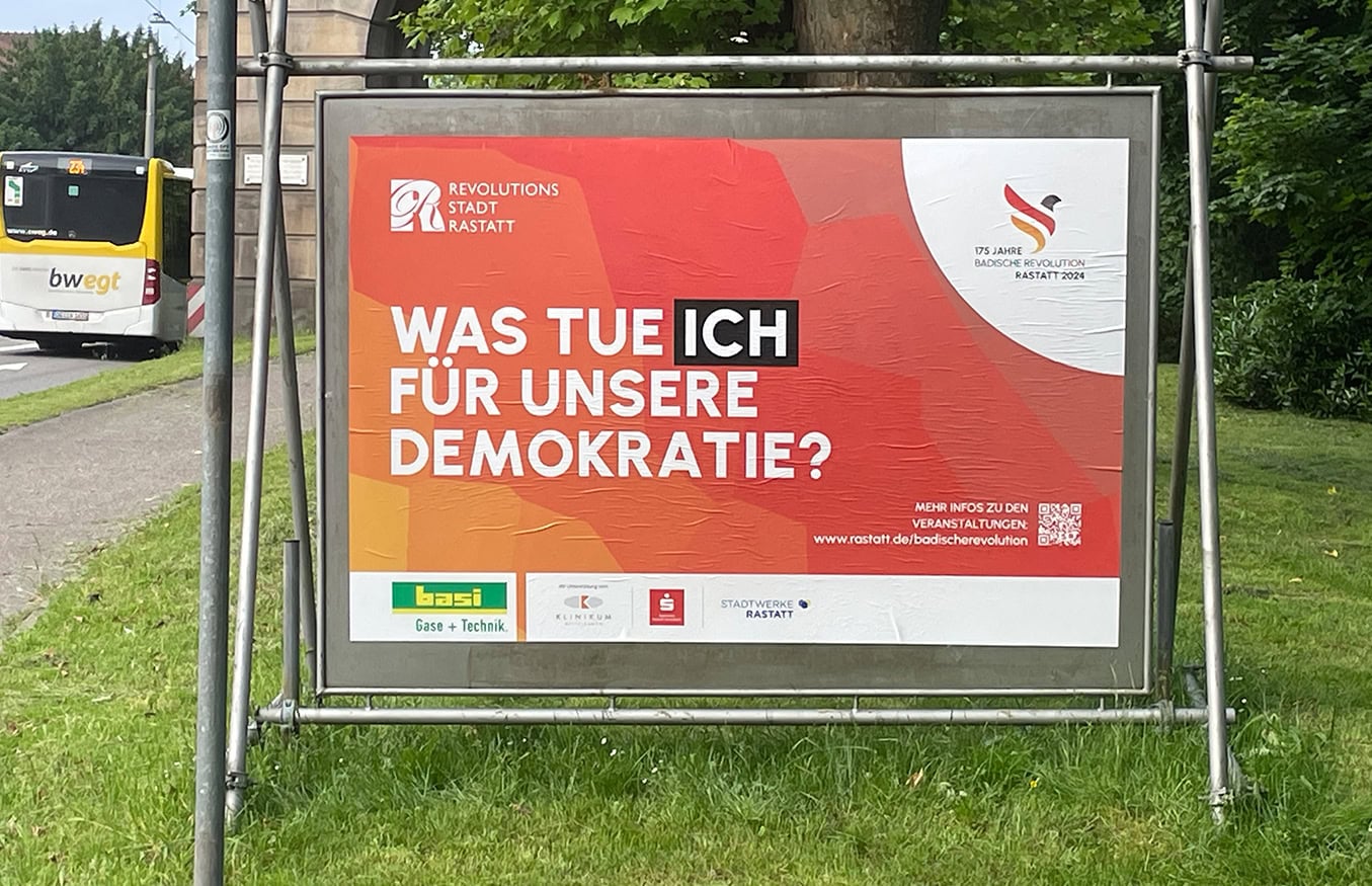 Plakatwand mit Text „WAS TUE ICH FÜR UNSERE DEMOKRATIE?“ und Logos von Sponsoren und ein QR-Code im Außenbereich.