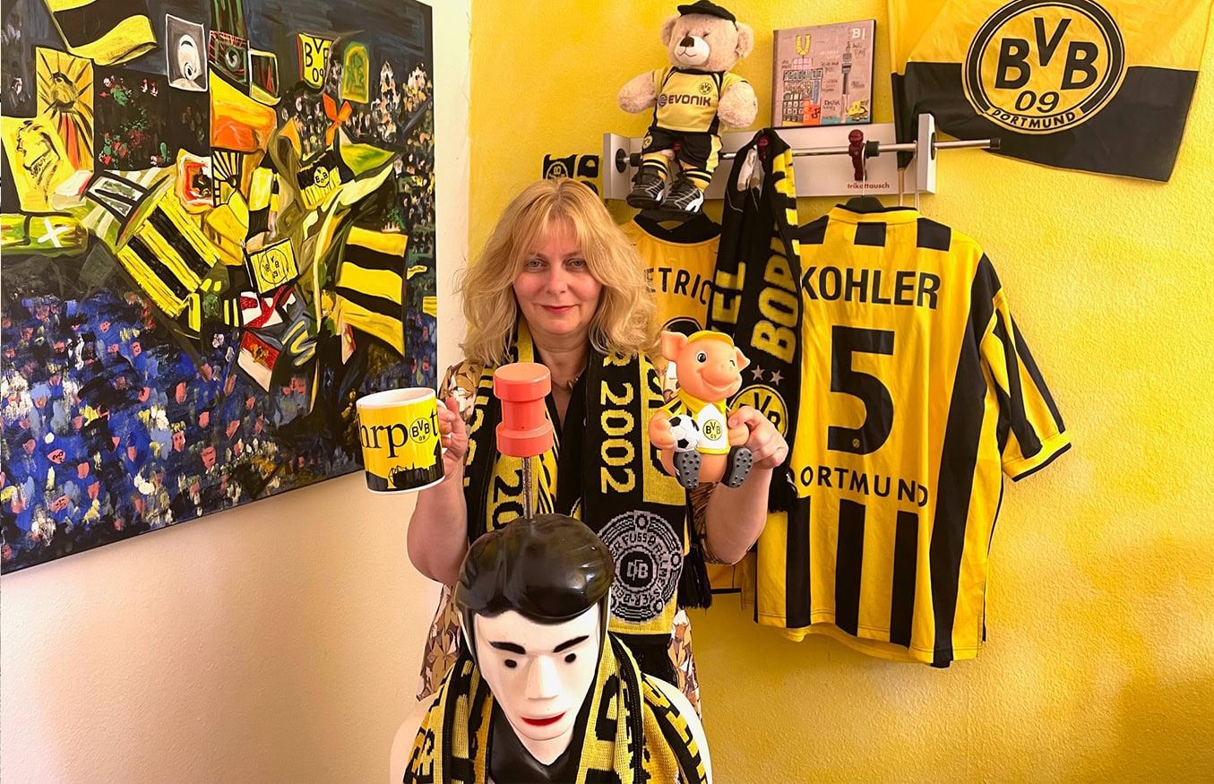 Frau umgeben von Dortmunder Fußball-Erinnerungsstücken, darunter Trikots, Schals, Poster und verschiedene Sammlerstücke.