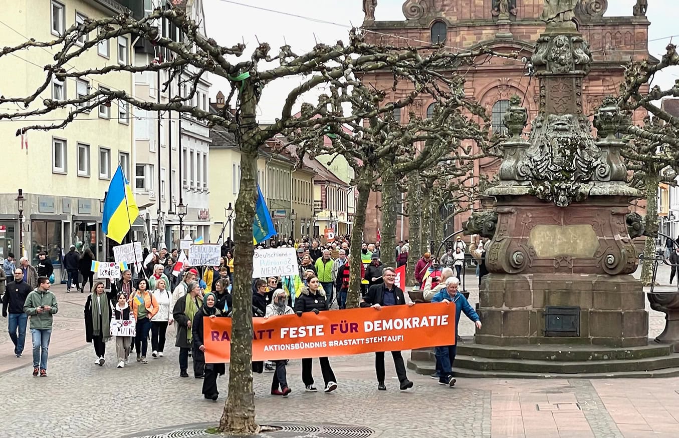 Eine Gruppe von Menschen marschiert mit Schildern, Flaggen und einem Banner in deutscher Sprache durch eine Stadt und wirbt für die Demokratie.