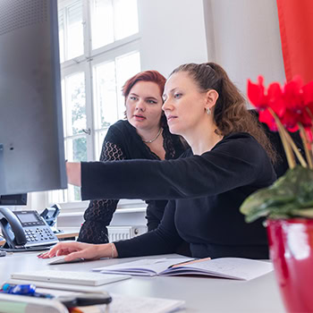 Zwei Frauen in einem Büro, eine zeigt auf einen Computerbildschirm, während die andere tippt. Im Vordergrund sind rote Blumen zu sehen.